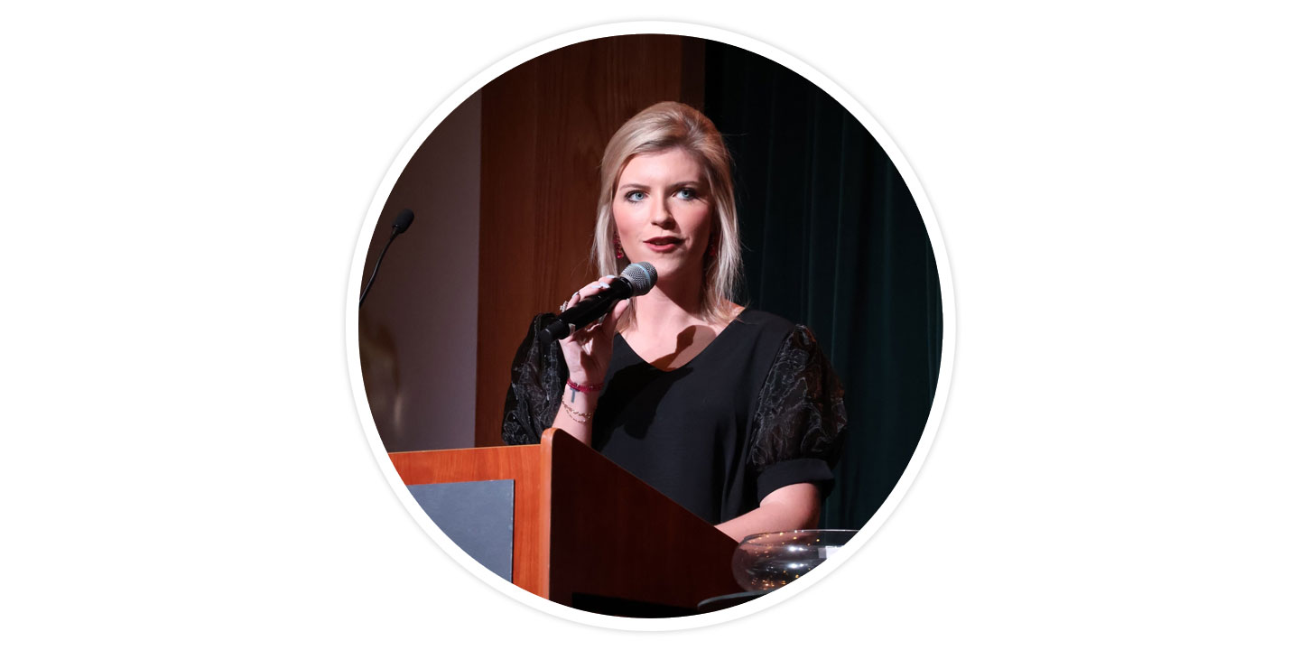 A woman with blonde hair holding a microphone and standing behind a podium talking, wearing a black shirt.